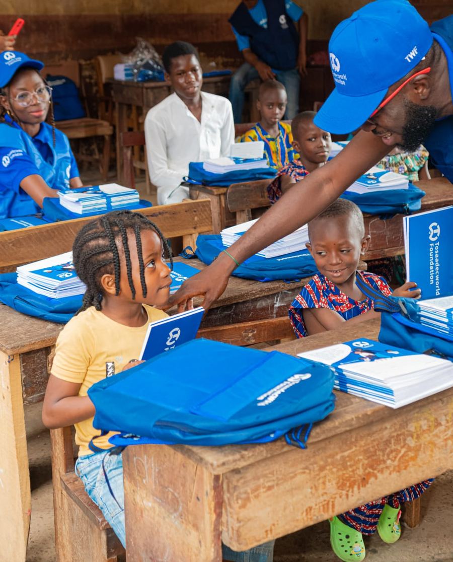 A TosaadecWorld Foundation volunteer hands a workbook to a schoolgirl at her desk during a school-essentials distribution.